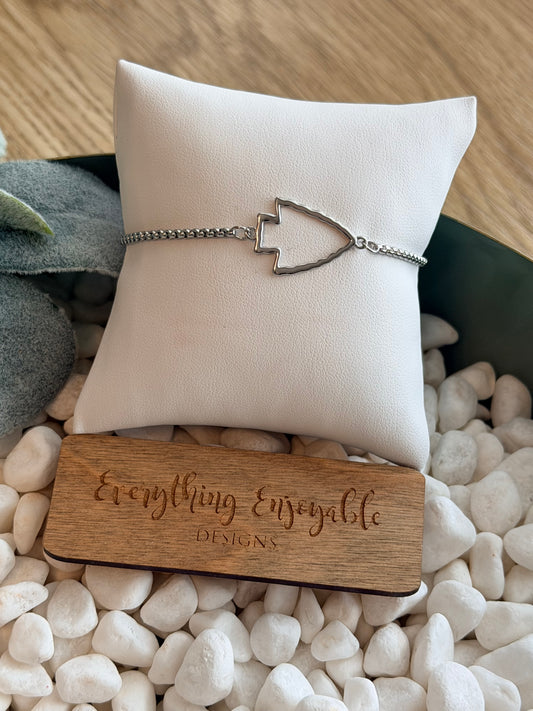Silver arrowhead bracelet on a white cushion with a wooden block labeled on pebbles.