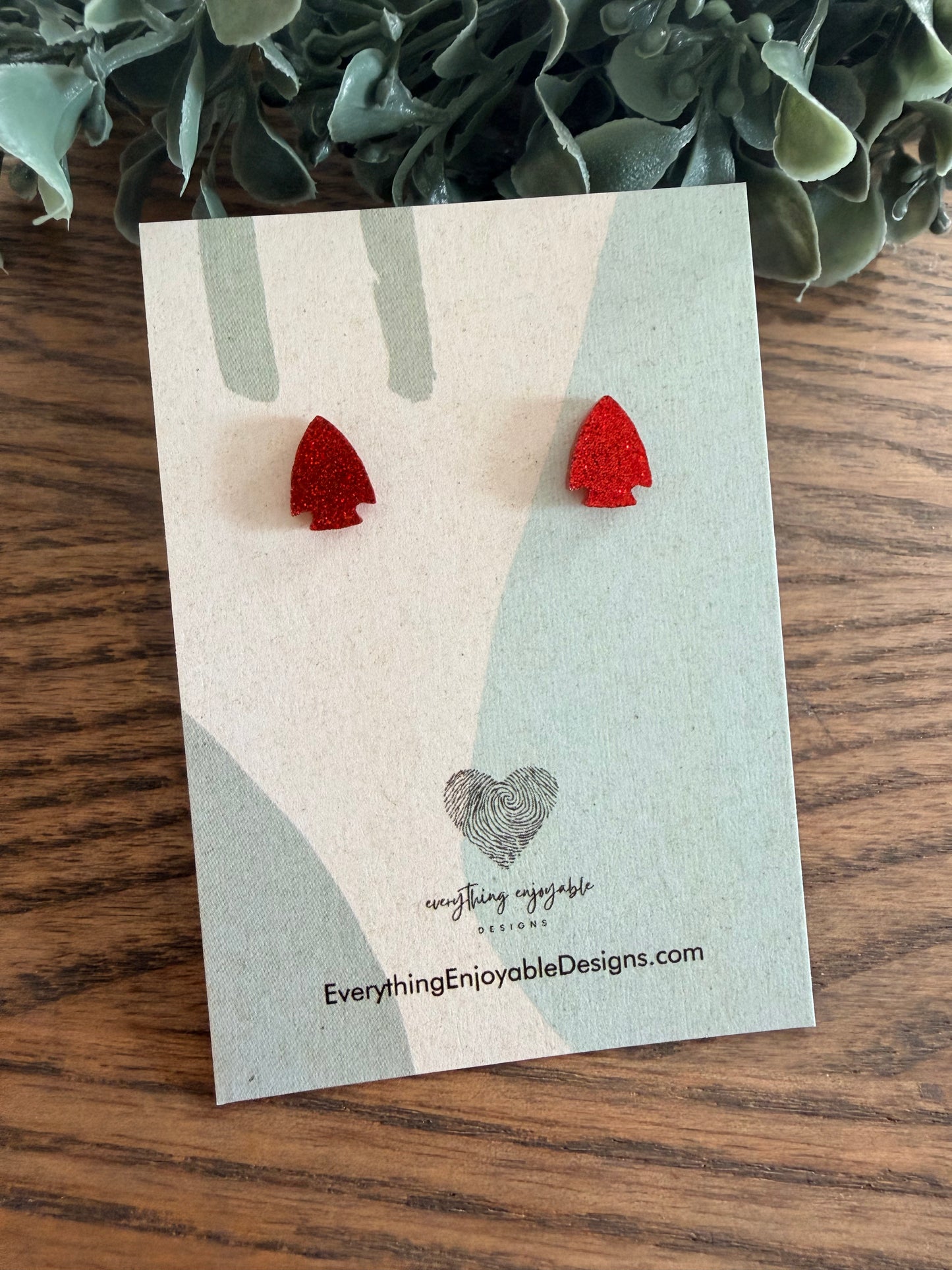Red glitter acrylic arrowhead earrings on a branded card with a wooden surface and plant in the background