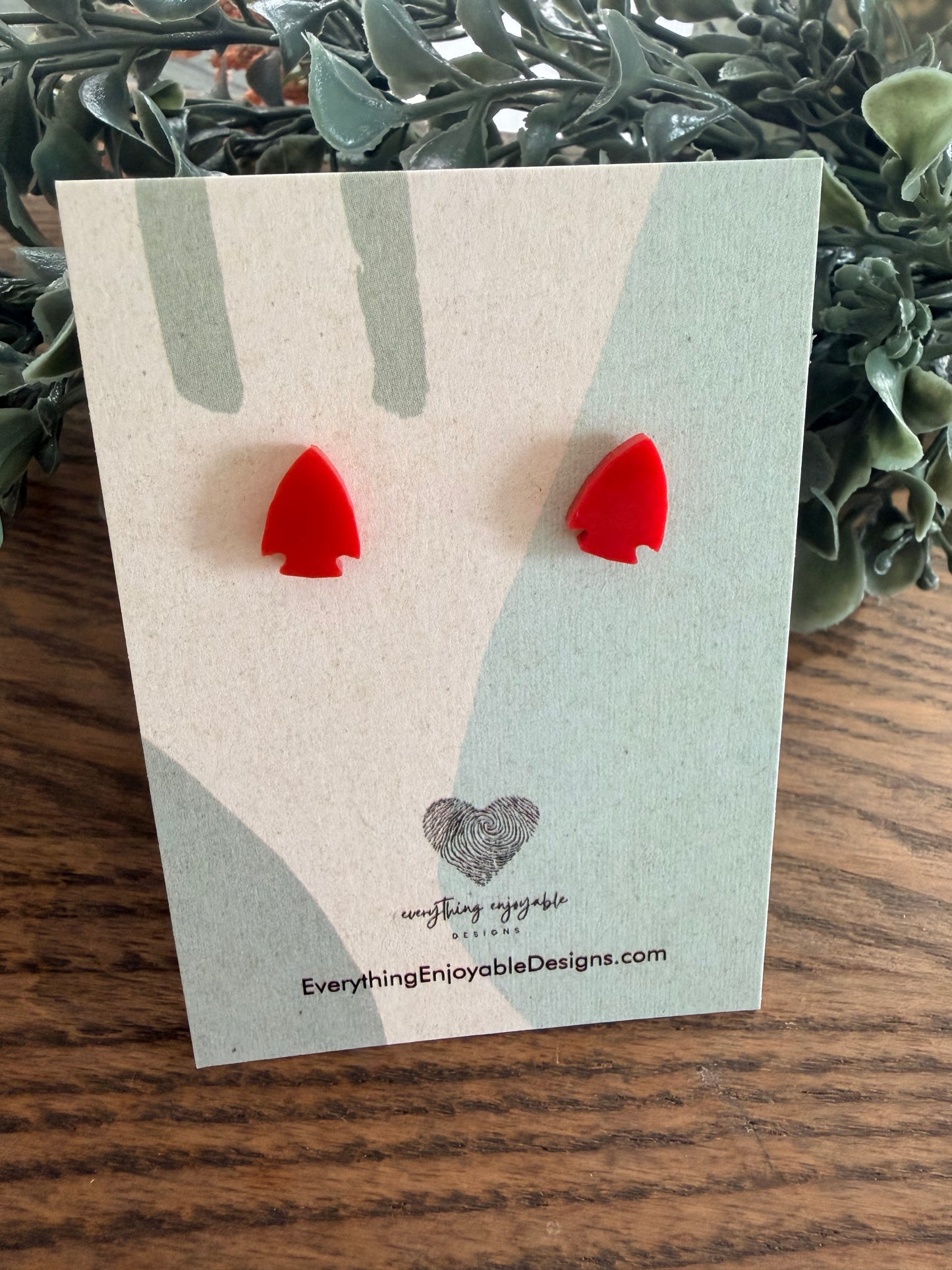 Red acrylic arrowhead earrings on a branded card with a wooden surface and plant in the background